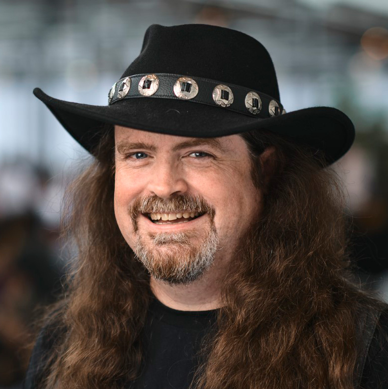 A photograph of Dylan Beattie, a bearded white man wearing a big stupid cowboy hat and grinning.