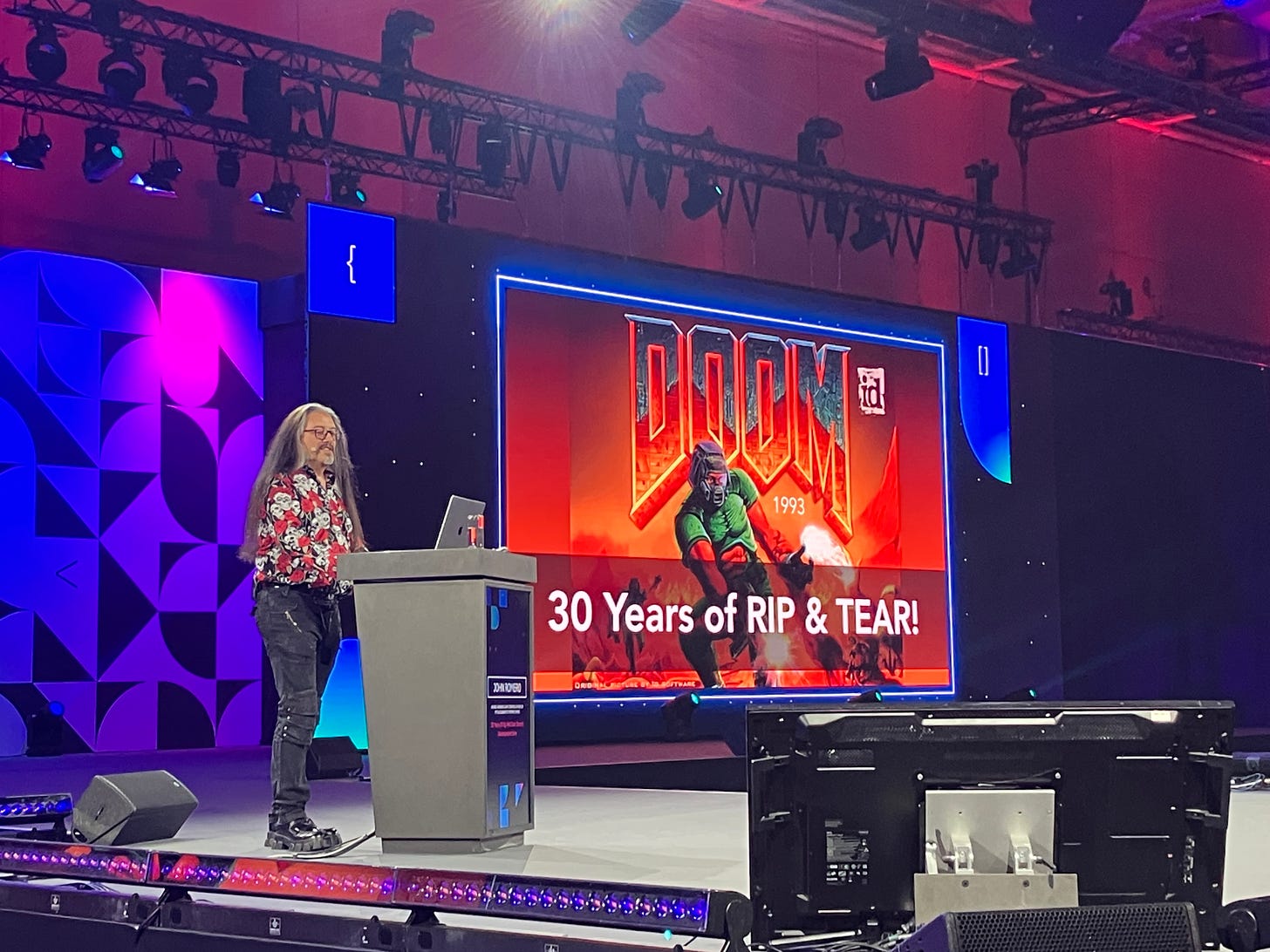 John Romero on stage, in front of a slide showing DOOM: 30 Years of RIP and TEAR. John wears a red and white floral shirt, black jeans, New Rock boots. He looks fabulous. 
