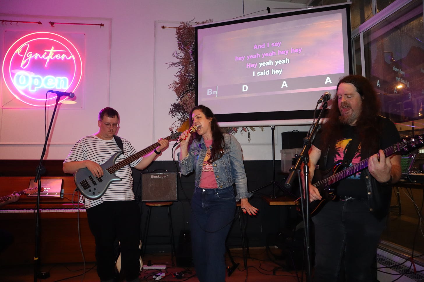 A neon sign reads "Ignition Open"; three musicians are playing and singing underneath a projector screen showing the song lyrics and chords. Everybody is very sweaty and there's cables and guitars everywhere. It looks like fun.