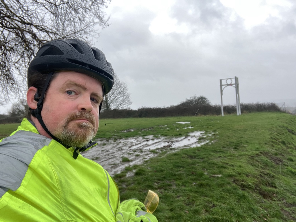 a sweaty man in a high-vis vest and cycle helmet at the top of a very muddy hill.