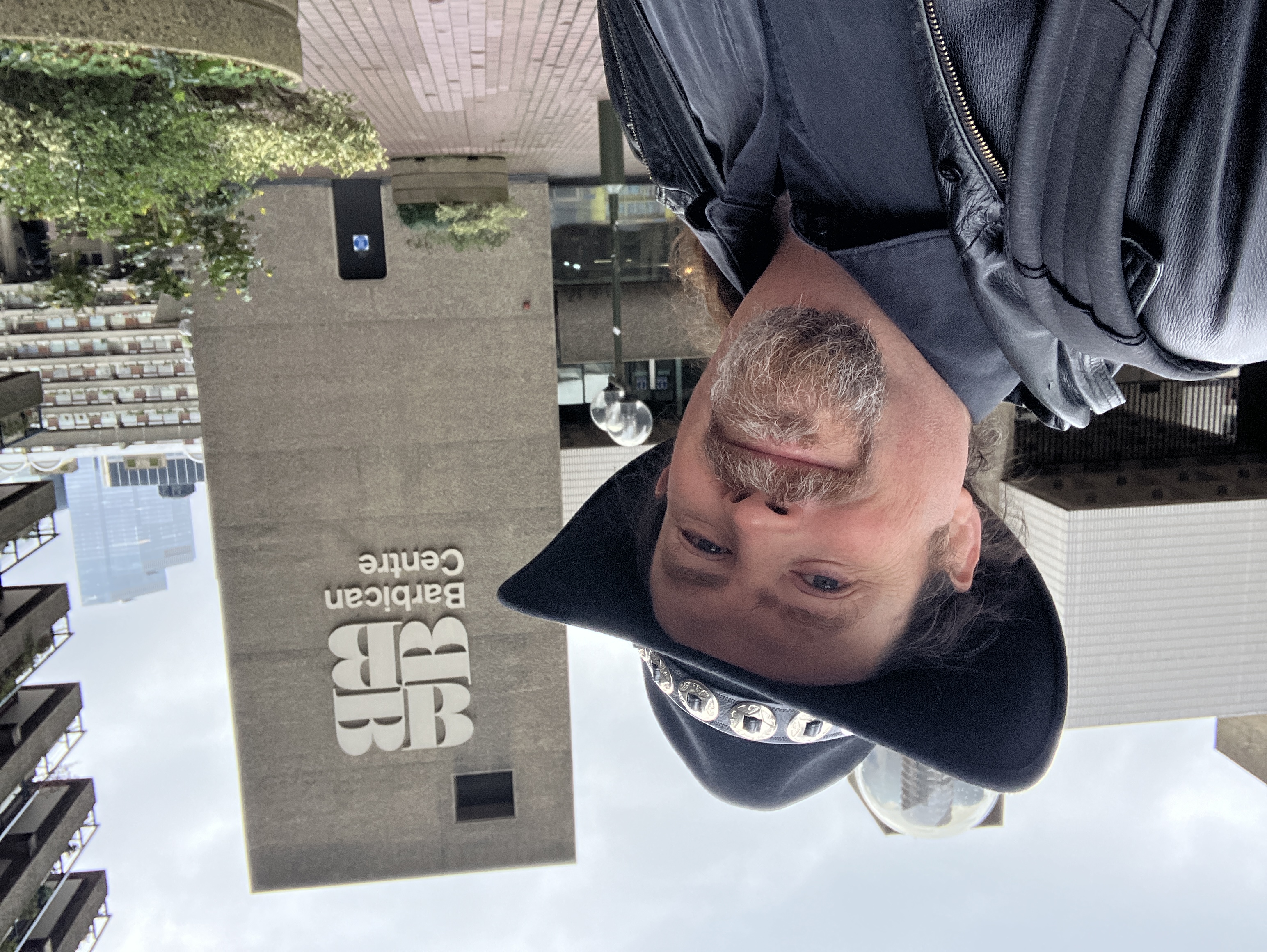A beardy man in a silly hat standing in front of about a million tons of existentialist concrete.